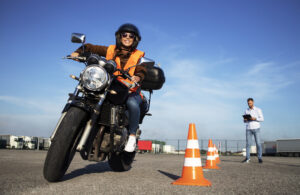 Kask takmış bir kadın öğrenci, motosiklet dersleri alıyor ve sürüş pratiği yapıyor. Arka planda trafik konileri ve bir kontrol listesiyle birlikte sürüşü değerlendiren bir eğitmen bulunuyor. Motosiklet sürüş okulu.
